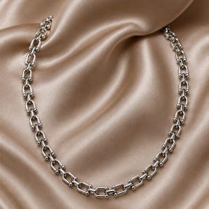 May include: A silver-toned necklace featuring a series of interconnected U-shaped links. Each link is joined by small, polished spherical beads at the connection points, creating a uniform chain design that forms a circular shape.