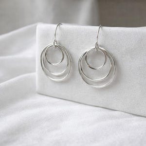 Op de afbeelding: Een paar zilverkleurige oorringen. Elke oorring heeft drie in elkaar geneste cirkelvormige ringen van afnemende grootte, opgehangen aan een eenvoudige haak. De oorbellen worden gepresenteerd tegen een witte achtergrond, wat hun minimalistische ontwerp benadrukt.