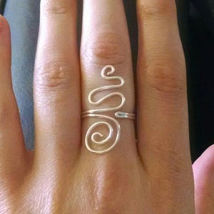May include: A silver-colored wire ring with a unique, abstract design. The ring features a spiral and a wavy line, creating a modern and artistic look. The ring is worn on a finger, showcasing its delicate craftsmanship.