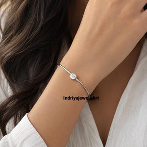 Puede incluir: Una pulsera plateada con un pequeño diseño de espiral blanco. La pulsera es fina y delicada, y la espiral está centrada en la muñeca. La imagen también incluye el texto "IndriyajewelsArt".