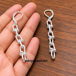 May include: A pair of silver-toned drop earrings featuring a vertical chain design with U-shaped links and small spherical accents at each joint. Each earring is finished with a secure lever-back closure mechanism for attachment to the earlobe.