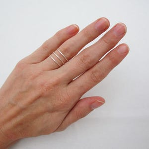 May include: A silver ring with three thin bands is worn on a finger. The ring is simple and minimalist in design. The hand is positioned against a white background.