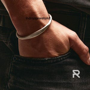 以下が含まれることがあります： 曲線的なデザインのシルバーカラーのカフブレスレット。ブレスレットは手首に着用されており、画像には「IndriyajewelsArt」の文字が含まれています。カフはシンプルでエレガントなジュエリーです。