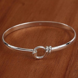 May include: A polished silver bangle bracelet featuring a hook and eye clasp. The bracelet's design is simple and elegant, with the clasp as a key detail. The metal is highly reflective, catching the light.