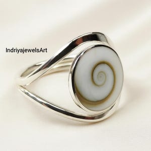 Puede incluir: Anillo de plata con una piedra blanca redonda y grande con un diseño en espiral. El anillo tiene un diseño abierto y moderno con un acabado pulido. El texto "IndriyajewelsArt" es visible en la imagen.