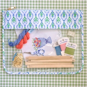 May include: A clear zippered pouch with a blue and green floral pattern trim, containing cross-stitch supplies. Includes embroidery floss, a needlepoint canvas with a floral design, scissors, wooden frame pieces, and gold-colored pins.