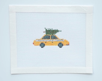 May include: A cross-stitch design featuring a yellow taxi cab with a green Christmas tree on top. The taxi has black and white checkered details and black wheels. The artwork is set against a light blue background and framed with a white border.