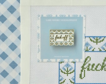 Op de afbeelding: Een rechthoekige naaldmagneet met een witte achtergrond, een blauwe en groene bloemenrand en de tekst "fuck off" in cursief schrift. De naaldmagneet is bevestigd op een lichtblauwe kaart met het opschrift "LAKE SHORE NEEDLEPOINT" en "NEEDLE MINDER", geplaatst op een witte rasterstof met een blauw en groen kruissteekpatroon.