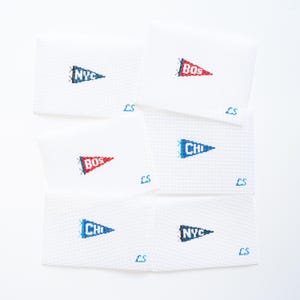 May include: A collection of white needlepoint pennants, each featuring a city abbreviation in a contrasting color. The pennants display "NYC", "BOS", and "CHI" in red and blue. The initials "LS" are stitched in blue on each pennant.