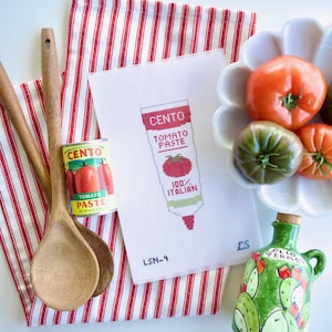 May include: A still life featuring a can of Cento tomato paste, a cross-stitch design of a tomato paste tube, wooden spoons, a decorative olive oil bottle, and a bowl of tomatoes. The cross-stitch reads "Cento Tomato Paste 100% Italian".