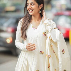 May include: A cream-colored, long-sleeved dress with floral embroidery. The dress has a V-neck and pleated skirt. A matching shawl with floral embroidery is draped over the arm. The woman is wearing silver earrings.