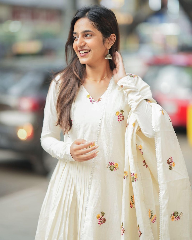 May include: A cream-colored, long-sleeved dress with floral embroidery. The dress has a V-neck and pleated skirt. A matching shawl with floral embroidery is draped over the arm. The woman is wearing silver earrings.