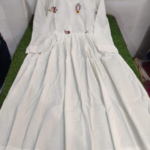 May include: A white long-sleeve dress with a V-neck and floral embroidery. The dress features a pleated skirt and a fitted bodice. The embroidery includes red, yellow, and green colors. The dress is hanging on a wooden hanger.