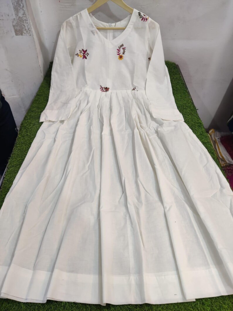 May include: A white long-sleeve dress with a V-neck and floral embroidery. The dress features a pleated skirt and a fitted bodice. The embroidery includes red, yellow, and green colors. The dress is hanging on a wooden hanger.