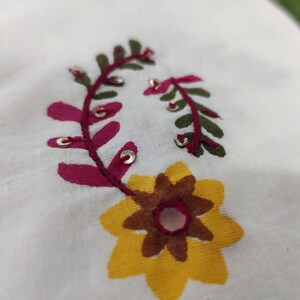May include: Close-up of white fabric with embroidered floral design. The embroidery features a yellow and brown flower, and burgundy and green leaves with silver accents. The fabric has a slightly textured appearance.