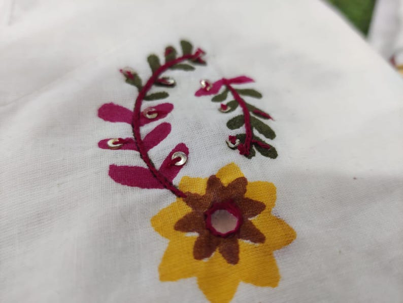 May include: Close-up of white fabric with embroidered floral design. The embroidery features a yellow and brown flower, and burgundy and green leaves with silver accents. The fabric has a slightly textured appearance.