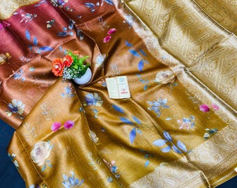 May include: A luxurious, floral-patterned saree in shades of brown and gold. The fabric features a gradient effect, transitioning from a deep burgundy to a golden hue. Delicate blue and pink floral designs adorn the saree, with a detailed gold border.