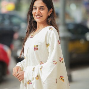 May include: A woman wearing a white embroidered outfit with long sleeves and a V-neck. The outfit features floral embroidery in shades of pink, yellow, and green. She is also wearing earrings and a necklace.