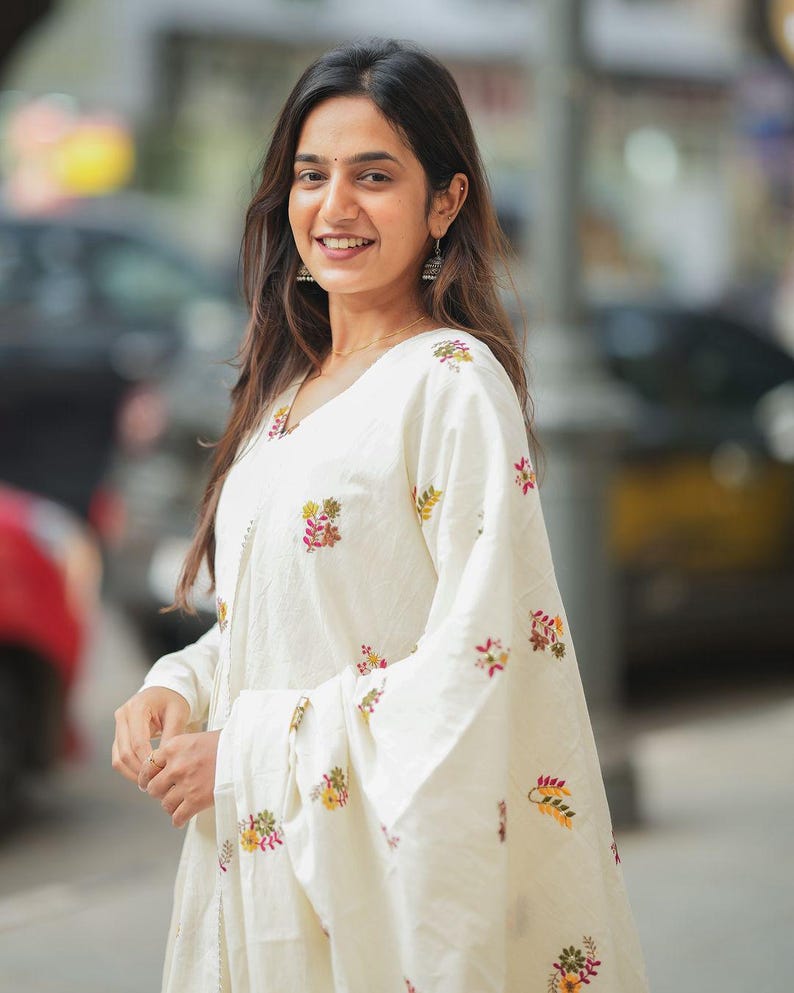 May include: A woman wearing a white embroidered outfit with long sleeves and a V-neck. The outfit features floral embroidery in shades of pink, yellow, and green. She is also wearing earrings and a necklace.