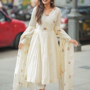 May include: Off-white, long-sleeved dress with a pleated skirt and floral embroidery. The dress has a V-neck and is paired with matching pants and a shawl. The outfit is worn with sandals.