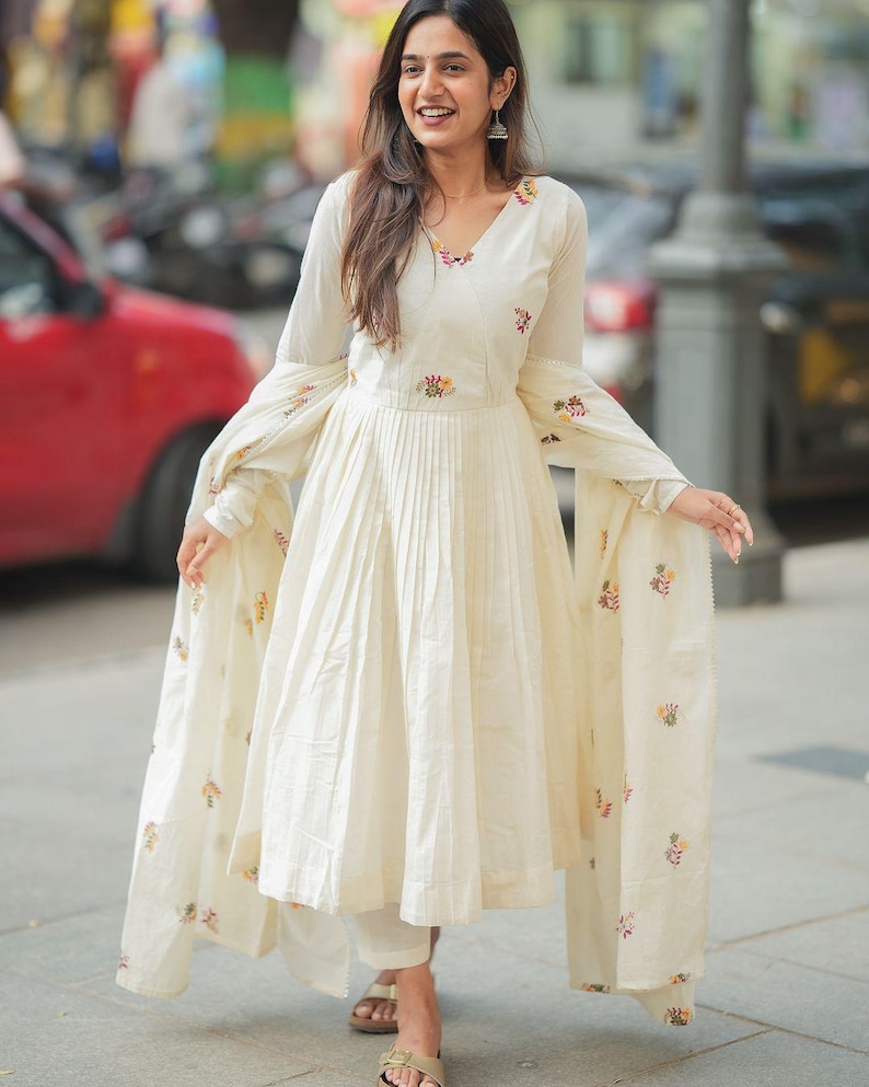 May include: Off-white, long-sleeved dress with a pleated skirt and floral embroidery. The dress has a V-neck and is paired with matching pants and a shawl. The outfit is worn with sandals.