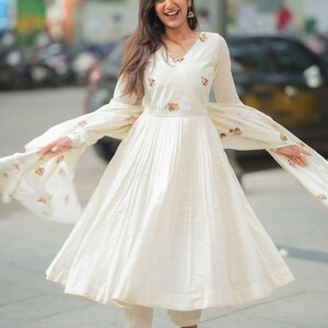 May include: Off-white, long-sleeved dress with a V-neck and pleated skirt. The dress features floral embroidery on the bodice and sleeves. The outfit includes matching pants and a flowing scarf, creating a coordinated ensemble.