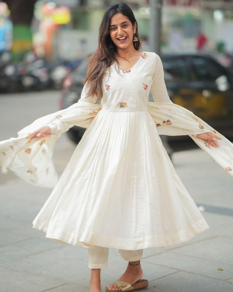 May include: Off-white, long-sleeved dress with a V-neck and pleated skirt. The dress features floral embroidery on the bodice and sleeves. The outfit includes matching pants and a flowing scarf, creating a coordinated ensemble.