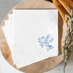 May include: A stack of white square paper napkins featuring a blue ink illustration of a champagne coupe glass with two cherubs and the text A+R and 12.24.20XX. The napkins have a textured diamond-patterned border along the bottom edge.