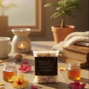 以下が含まれることがあります： 「Wicks & Wheel」の高級アロマのパッケージ、蜂蜜の瓶、ハイビスカスの花をあしらった静物画。白いセラミックのアロマウォーマーにはろうそくが灯っている。木製の表面に、背景には小さな鉢植えの植物が置かれている。