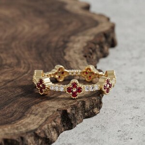 Puede incluir: Un anillo de oro con un patrón repetitivo de motivos florales de cuatro pétalos. Cada flor está engastada con cuatro pequeñas piedras rojas y una cuenta dorada central, alternando con secciones de tres piedras blancas claras en el aro.