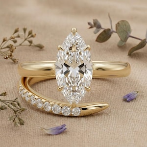 Puede incluir: Un anillo de compromiso de diamantes con corte marquesa y banda dorada. El anillo presenta un diamante grande y transparente y se combina con una banda dorada curva con diamantes más pequeños. El fondo es de color beige neutro.