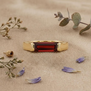 Könnte beinhalten: Ein goldener Ring mit einem rechteckigen, tiefroten Edelstein. Der Ring liegt auf einer neutralen Oberfläche, umgeben von getrockneten Blumen und lila Blütenblättern. Der Edelstein ist der Mittelpunkt und zeigt eine satte, lebendige Farbe.
