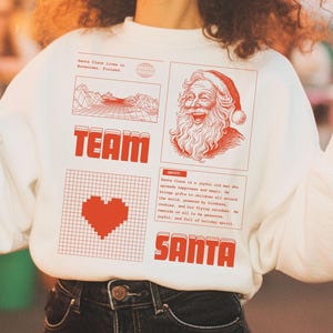Op de afbeelding: Een witte sweatshirt met een rood grafisch ontwerp met de woorden "TEAM SANTA", een pixelhart en een illustratie van de kerstman. De persoon draagt twee drankjes met rietjes.