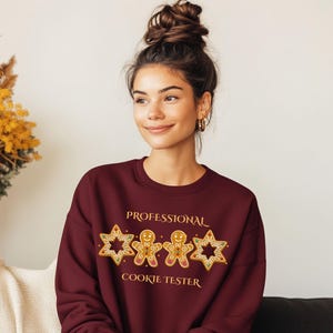 Op de afbeelding: Een bordeauxrode sweatshirt met de tekst "PROFESSIONAL COOKIE TESTER" in gouden letters. Het ontwerp bevat peperkoekmannetjes en stervormige koekjes. Een feestelijk kledingstuk voor de feestdagen.