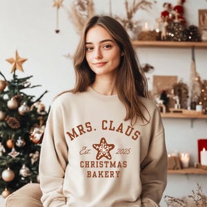 Op de afbeelding: Een beige sweatshirt met de tekst "MRS. CLAUS CHRISTMAS BAKERY Est. 2025" in bruine letters. Het ontwerp bevat een stervormige peperkoek-afbeelding. Het sweatshirt maakt deel uit van een collectie met kerstthema.