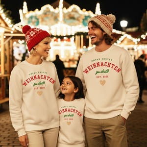 Op de afbeelding: Drie mensen dragen bijpassende witte sweatshirts met de Duitse tekst "UNSER ERSTES WEIHNACHTEN zu dritt EST. 2025" in rood en groen. Eén persoon draagt een rood-witte muts. Een gouden hartafbeelding staat ook op de sweatshirts. De foto is 's nachts genomen.