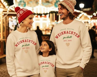 Puede incluir: Tres personas con sudaderas blancas a juego con el texto alemán "UNSER ERSTES WEIHNACHTEN zu dritt EST. 2025" en rojo y verde. Una persona lleva un gorro con estampado rojo y blanco. Un corazón dorado también está en las sudaderas. La imagen es de noche.