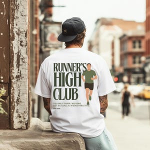 Pode incluir: Camiseta branca com o texto "RUNNER'S HIGH CLUB" em verde escuro. A camiseta apresenta um gráfico de uma figura correndo e a citação: "YOU MAY THINK I'M DYING, BUT ACTUALLY I'M ENJOYING THIS." ESTD 2008.