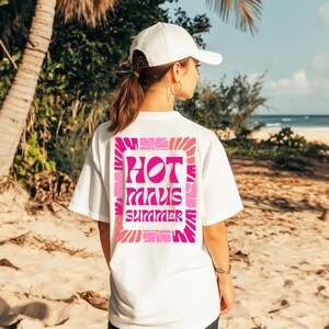 Puede incluir: Camiseta blanca con un gr&aacute;fico rosa y fucsia que dice "HOT MINUS SUMMER". El dise&ntilde;o est&aacute; enmarcado con un patr&oacute;n floral. La camiseta se usa en una playa con palmeras y el oc&eacute;ano de fondo.