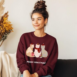 Op de afbeelding: Bordeaux sweatshirt met een feestelijk ontwerp met drie cartoonkatten die kerstmutsen en sjaals dragen. De katten houden een cadeau en ornamenten vast. De tekst "MERRY CHRISTMAS!" staat eronder gedrukt. Een kledingstuk met een vakantiethema.