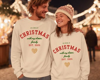Pode incluir: Duas pessoas vestindo moletons creme combinando com o texto "FIRST CHRISTMAS with my chosen family EST. 2025" em vermelho e verde. Um coração dourado está abaixo do texto. O fundo está desfocado com luzes de Natal.