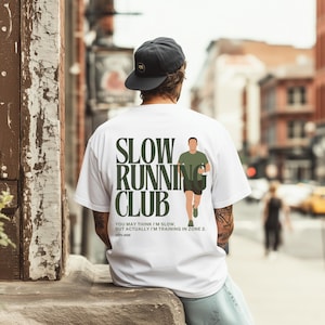 Könnte beinhalten: Weißes T-Shirt mit dem Text "SLOW RUNNING CLUB" in Dunkelgrün. Das Shirt zeigt eine Grafik eines Läufers und den Text "YOU MAY THINK I'M SLOW, BUT ACTUALLY I'M TRAINING IN ZONE 2." Die Person trägt eine schwarze Kappe.