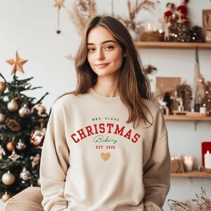 Op de afbeelding: Een beige sweatshirt met de tekst "MRS. CLAUS CHRISTMAS Bakery EST. 2025" in rood en groen, met een gouden hart. De persoon draagt de trui voor een kerstboom en decoratieve planken.