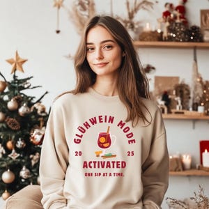 Op de afbeelding: Een beige sweatshirt met de tekst "Glühwein Mode Activated" in bordeauxrood. De afbeelding toont een mok glühwein, kaneelstokjes en sinaasappelschijfjes. De tekst "One sip at a time" staat eronder. Perfect voor de feestdagen.