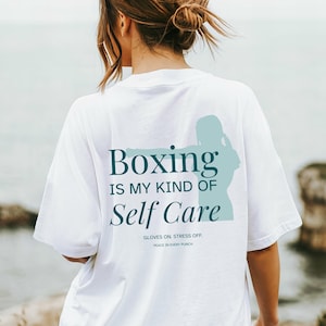 Puede incluir: Camiseta blanca con el texto "Boxing IS MY KIND OF Self Care" en color turquesa. Una silueta turquesa de un boxeador está a la derecha de la palabra "Boxing". El texto "GLOVES ON. STRESS OFF. PEACE IN EVERY PUNCH" está debajo.