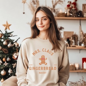 Op de afbeelding: Beige sweatshirt met de tekst "MRS. CLAUS GINGERBREAD BAKERY ESTD 2025" en een gingerbread man afbeelding. Het ontwerp is in warme tinten, perfect voor de feestdagen.