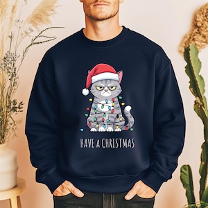 Op de afbeelding: Marineblauwe sweatshirt met een chagrijnige cartoonkat met een kerstmuts en omwikkeld met kerstverlichting. De tekst "HAVE A CHRISTMAS" staat eronder. Een feestelijk kledingstuk met een kerstthema.
