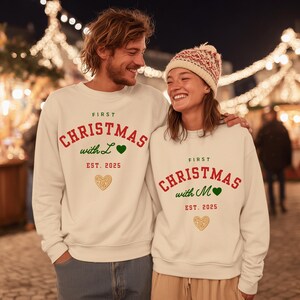 Op de afbeelding: Witte sweatshirts met de tekst "First Christmas" in rood, "with L" en "with M" in groen en "EST. 2025" in rood. Elk sweatshirt heeft een gouden hartje. Een lachend stel omhelst elkaar.