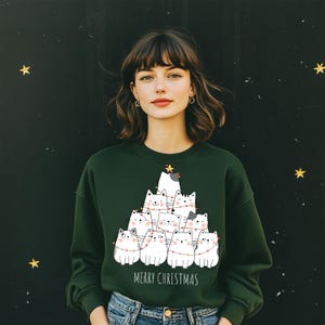 Op de afbeelding: Een donkergroene sweatshirt met een kerstboomontwerp van witte katten en lichtslingers. De woorden "MERRY CHRISTMAS" staan onder de katten gedrukt. De persoon draagt een blauwe spijkerbroek.