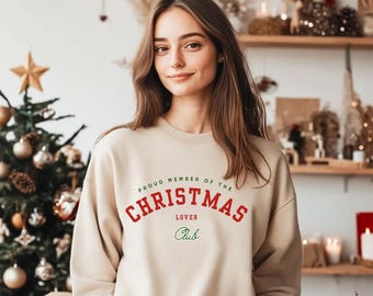 Pode incluir: Um moletom bege com o texto "PROUD MEMBER OF THE CHRISTMAS LOVER CLUB" em vermelho e verde. O moletom é de gola redonda e está sendo usado por uma pessoa. O fundo inclui uma árvore de Natal e prateleiras decorativas.